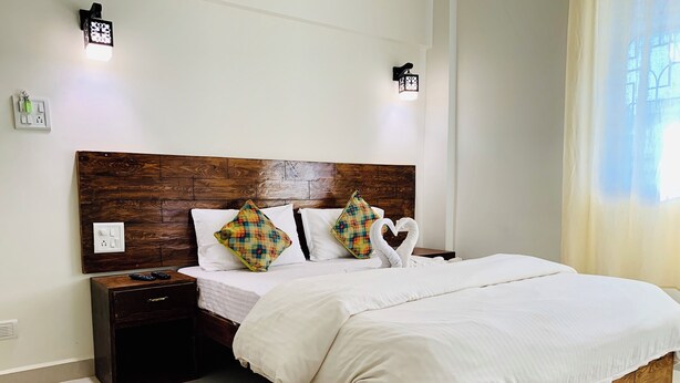 A neatly made bed with white linens, a wooden headboard, and two decorative pillows. There are bedside tables with remote controls and a wall socket. - Mamata Courtyard Anjuna (Anjuna)