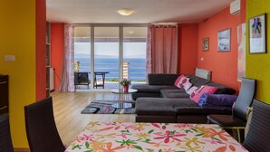 A living room with a sofa, chairs, a small table, and a view of the sea through a large sliding glass door. - Colourful Seaview Apartment (Kostrena)