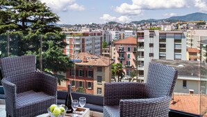 A balcony with wicker chairs and a table with a bottle and glasses, overlooking a cityscape with buildings and a mountain in the distance. - Panoramic Penthouse in Lugano (Lugano)