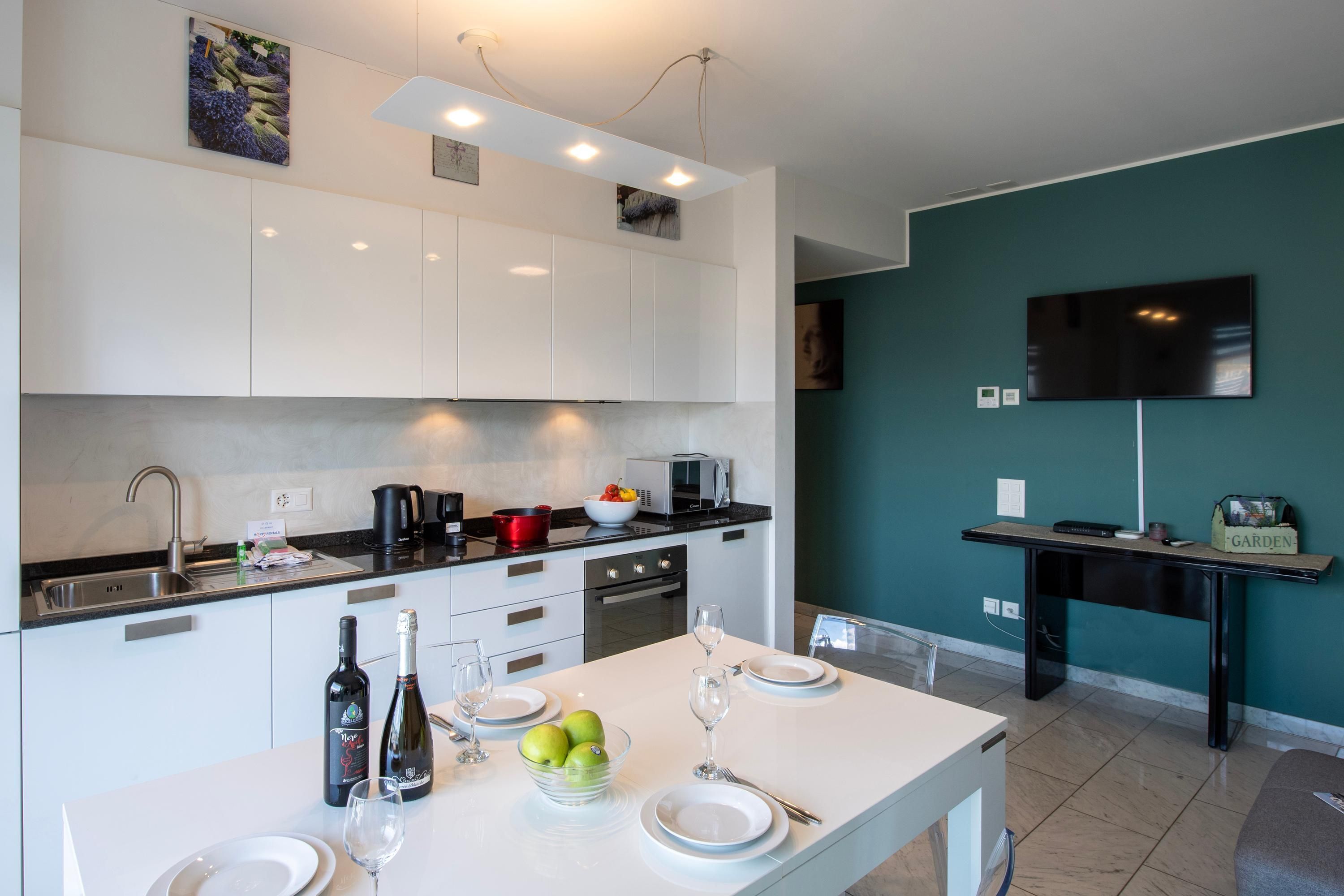 A modern kitchen with white cabinets, a dining table set for two, and a TV mounted on the wall.