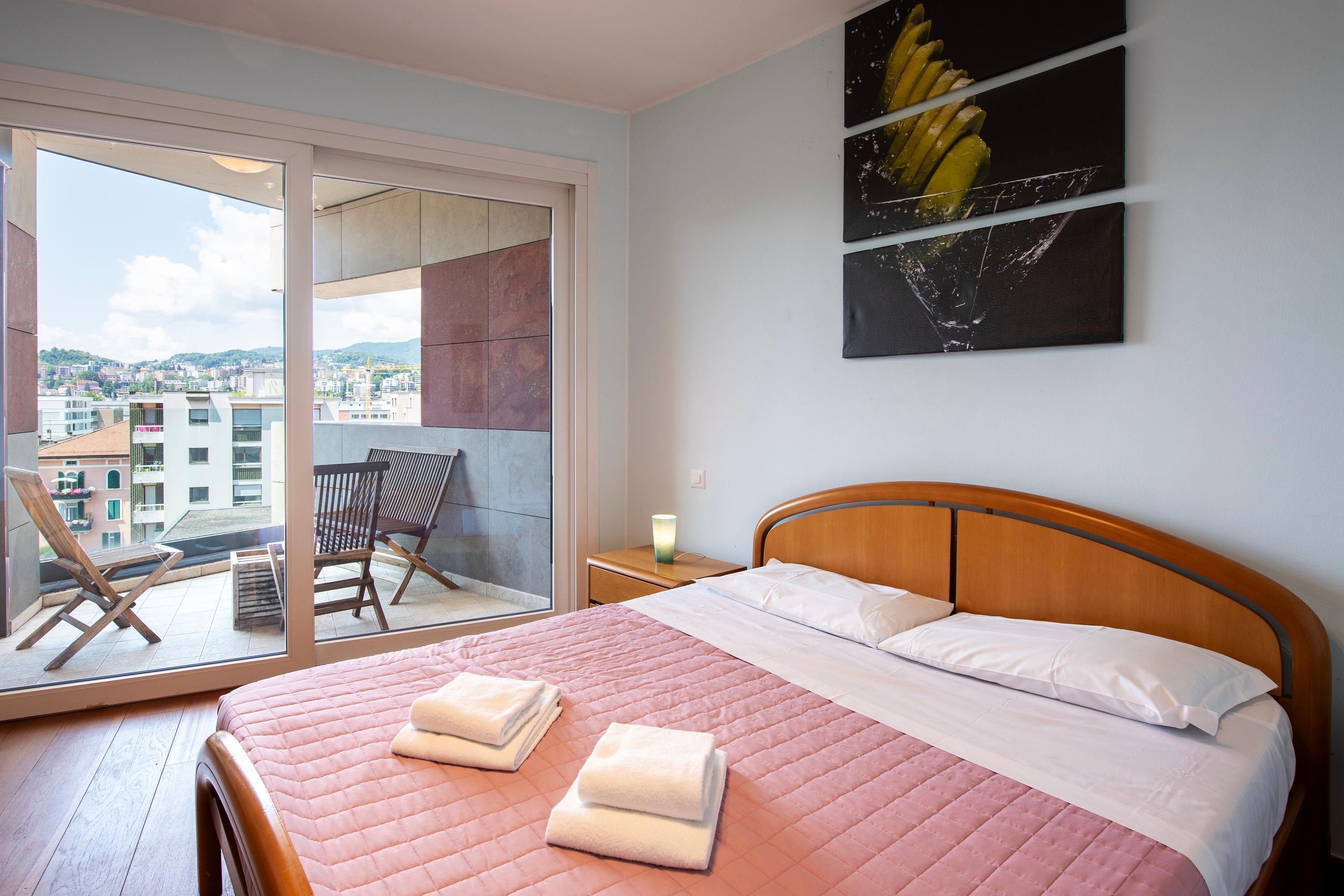 A bedroom with a bed, a window with a view of buildings, a chair, and a nightstand.