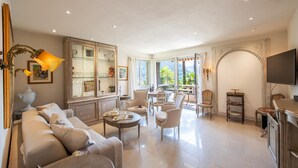 Ein geräumiges Wohnzimmer mit Sofa, Sesseln, einem Couchtisch und einem Fernseher. Es gibt einen Essbereich mit Tisch und Stühlen, sowie eine Küchenzeile mit Schränken und einem Herd.