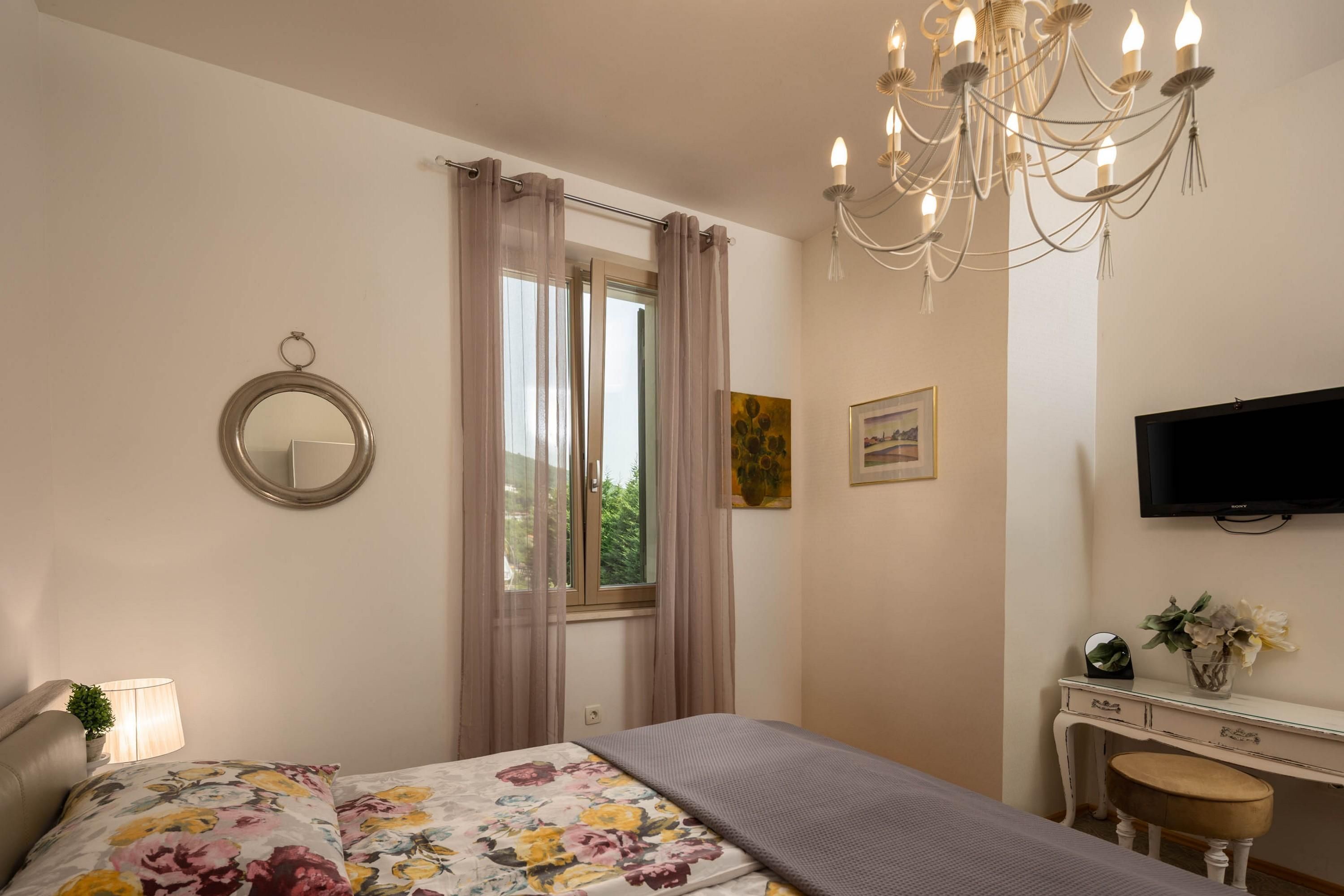A bedroom with a bed, a desk, a chair, a TV, a mirror, and a window with curtains.