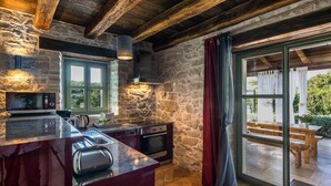 A rustic kitchen with stone walls, wooden beams, a large island with a built-in microwave, and a view to an outdoor patio. - Villa Zvonar in Karojba (Karojba)
