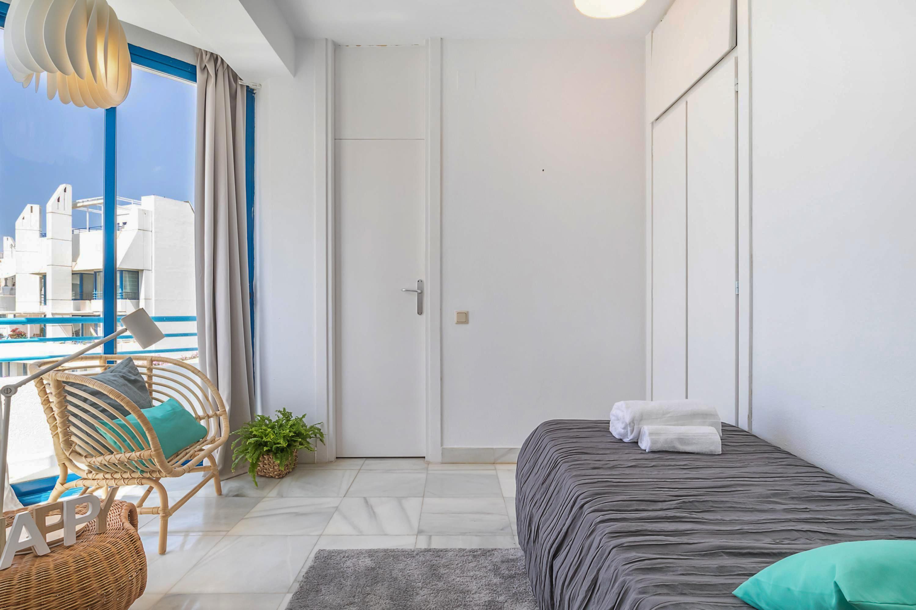 A modern hotel room with a bed, a chair, a small table, and a view of a pool and buildings.