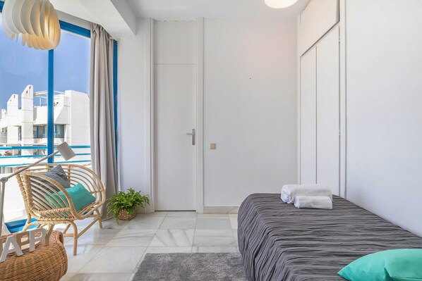 A modern hotel room with a bed, a chair, a small table, and a view of a pool and buildings.