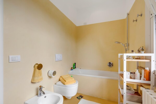 Un baño con inodoro, lavabo y bañera. Hay un estante con artículos de aseo y un perchero para toallas.