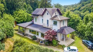 A two-story house with a sloped roof, surrounded by lush greenery and trees.