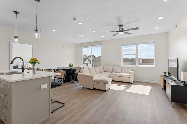 Relax in a spacious living area with a large sofa and dining table for four, surrounded by natural light and ceiling fan.