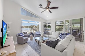 A spacious living room with a large flat-screen TV, a ceiling fan, and a view of the outdoors through large sliding glass doors.