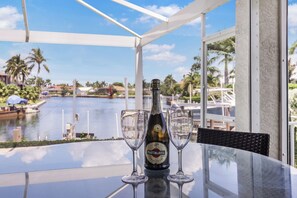 A table with a bottle of Martini Rosso wine and two glasses, overlooking a body of water with palm trees and buildings in the background.