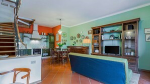 A spacious living area with a dining table, a sofa, and a TV. A kitchen with green cabinets and a wooden staircase is visible in the background.