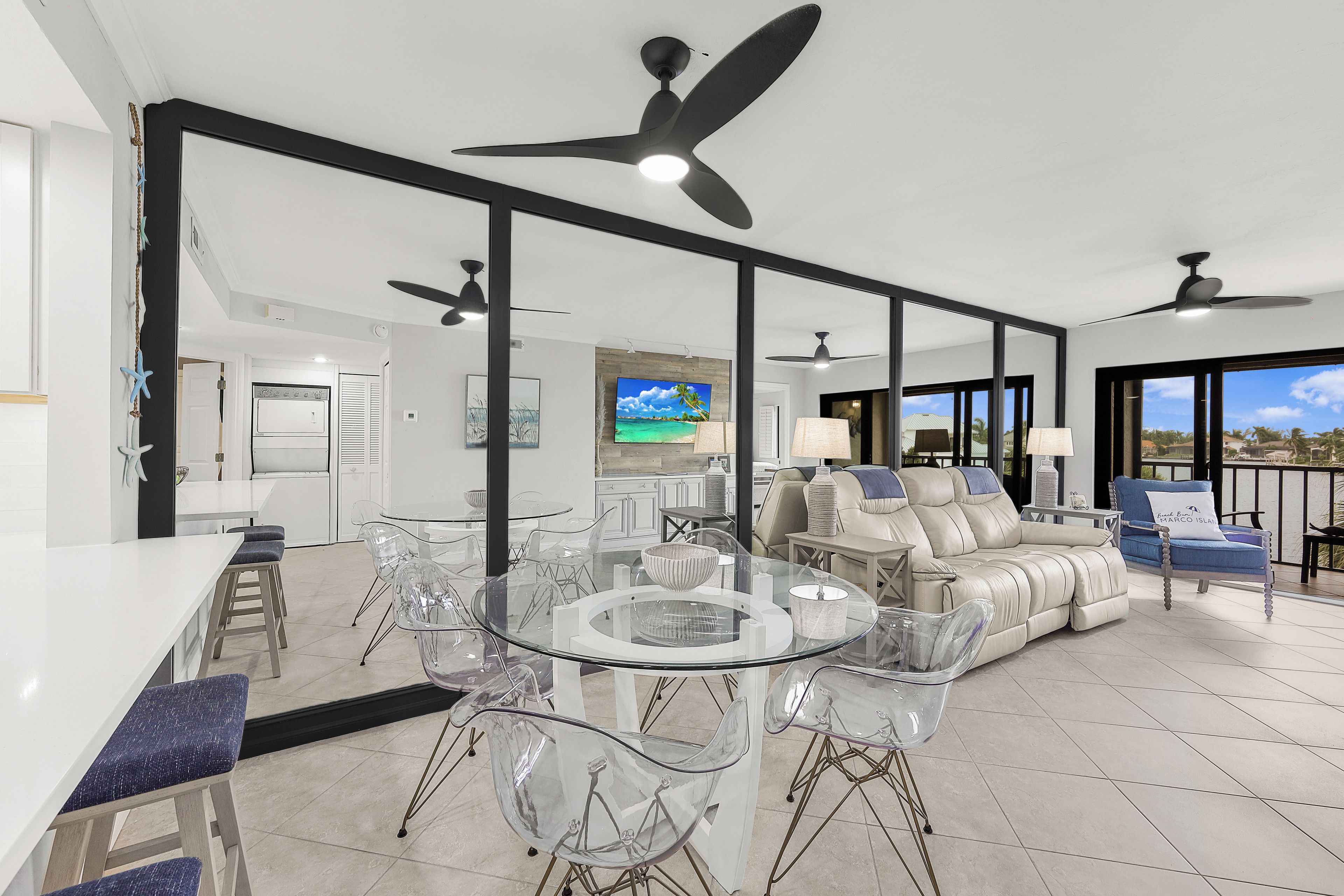 A spacious living area with a glass dining table, clear chairs, a leather sofa, and a balcony with a view.