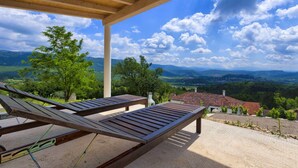 A covered outdoor seating area with two wooden lounge chairs, a view of a rural landscape with trees and a house, and a clear blue sky with clouds. - Villa Lapis in Buzet (Buzet)