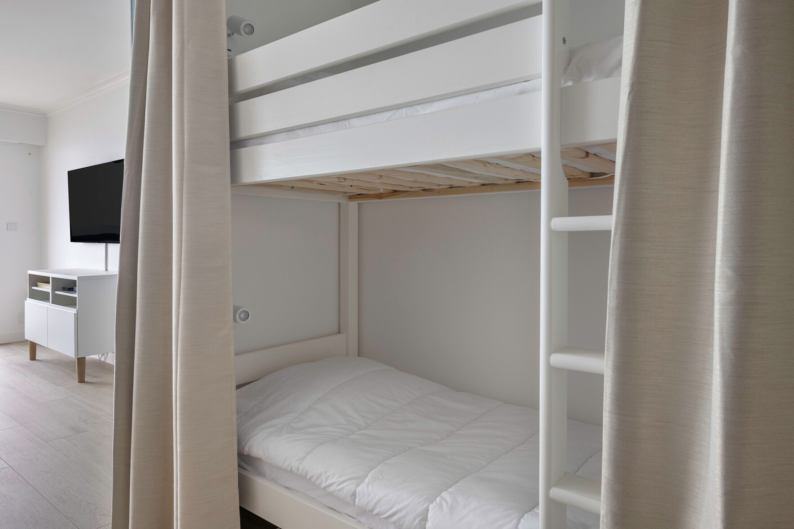Habitación con literas, un cabecero blanco, un televisor de pantalla plana montado en la pared y un aparador blanco con estantes abiertos.