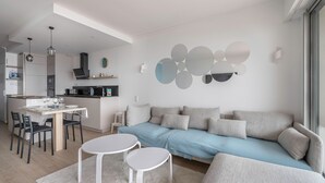 Una sala de estar moderna con un sofá, dos mesas blancas y una cocina al fondo.