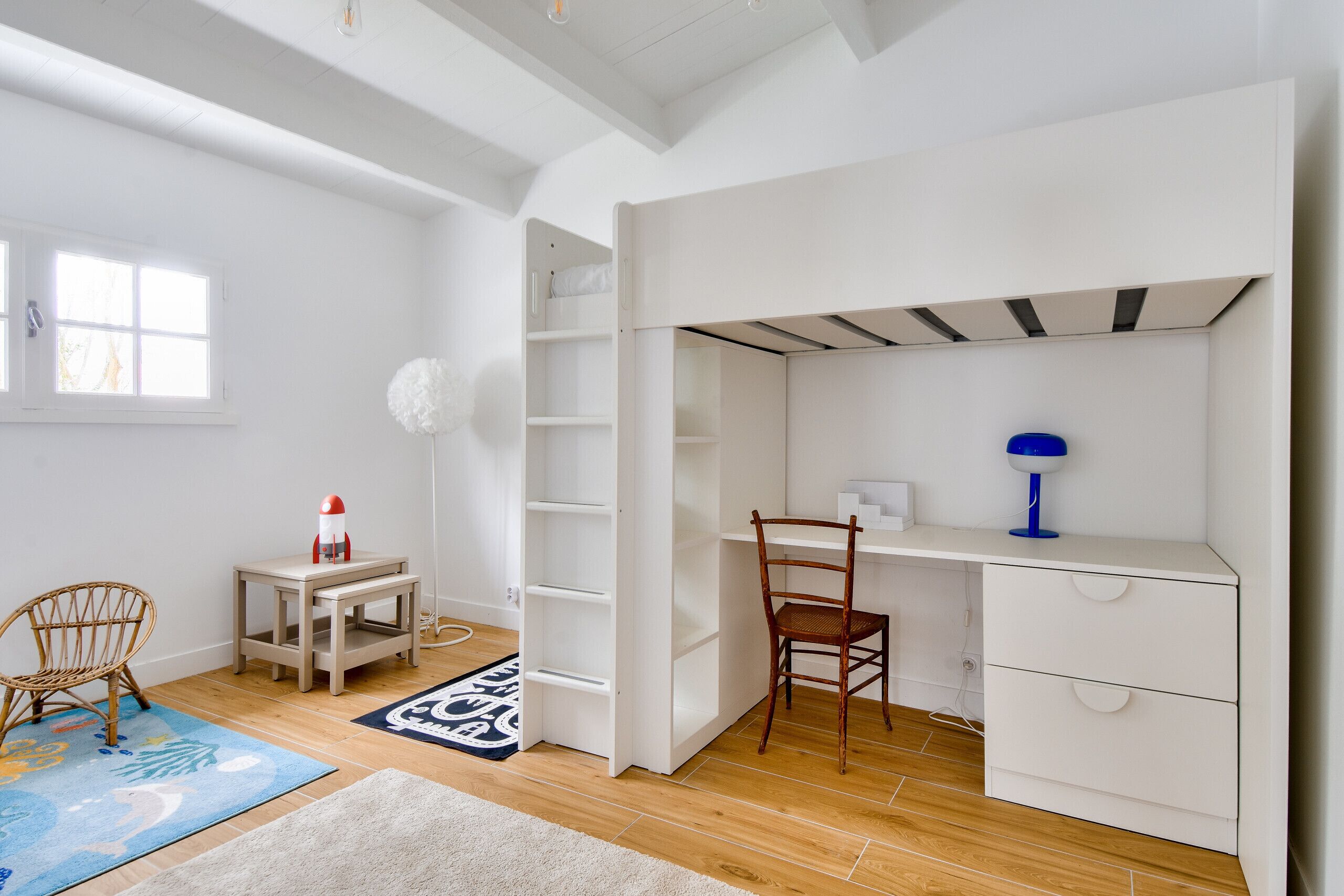 Una habitación con un escritorio blanco, una silla, una estantería y una ventana.