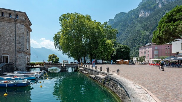 A riverside view with boats docked, a stone bridge, and buildings along the water. - Life View Luxury Apartment (Riva del Garda)