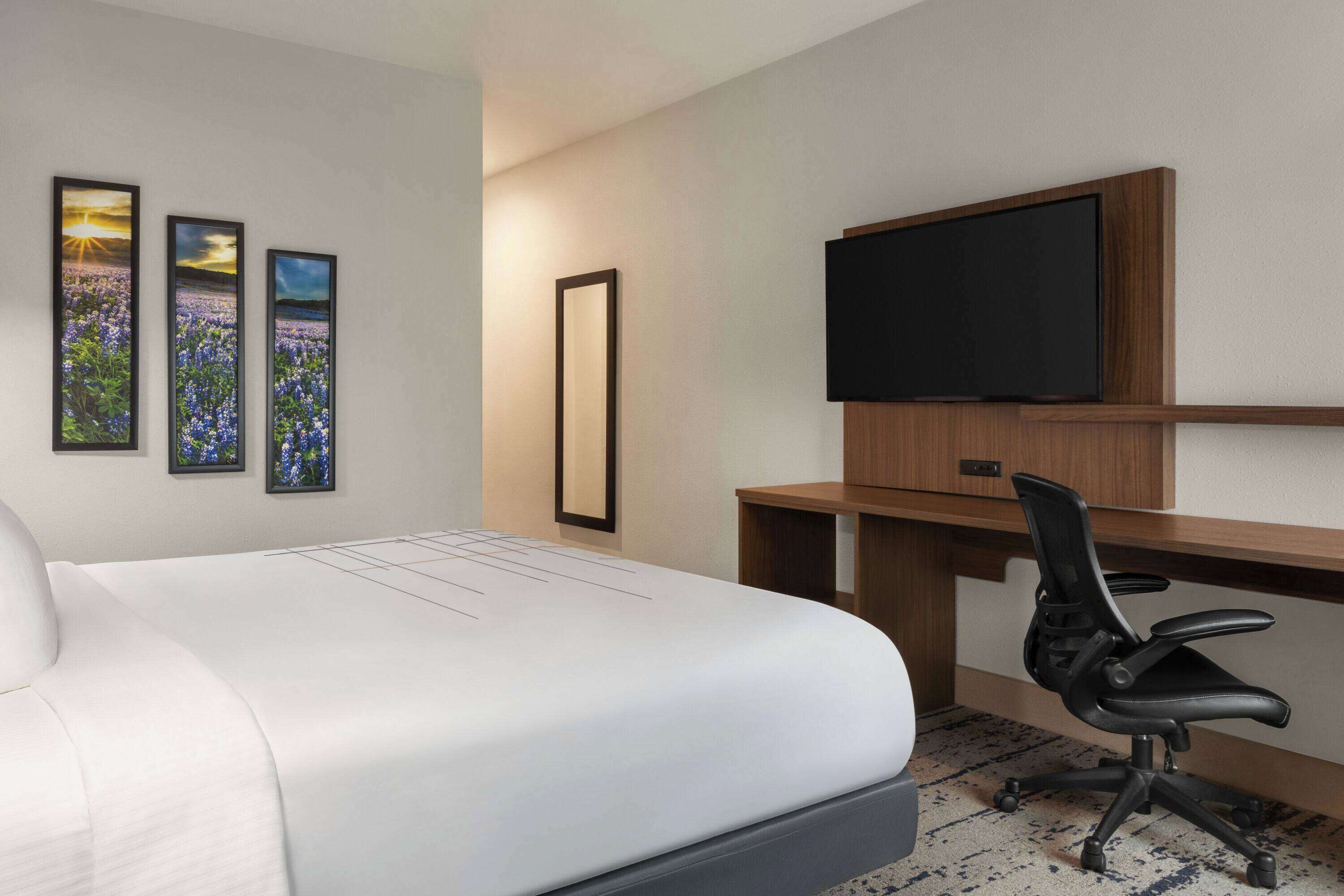 Une chambre d’hôtel avec un grand lit, un bureau avec une chaise, un téléviseur à écran plat et trois tableaux encadrés au mur.