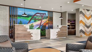 A reception area with a modern design, featuring a large digital screen displaying a scenic view, a staircase leading to a second floor, and a seating area with patterned sofas. - La Quinta Inn & Suites By Wyndham Austin South (Austin)
