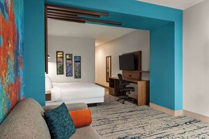 A hotel room with a bed, a desk with a computer, a TV, and a colorful abstract painting.