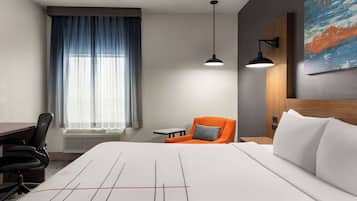 A hotel room with a large bed, a desk, an office chair, a small table, a chair with an orange cushion, and a painting on the wall.