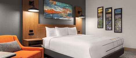A modern hotel room with a large bed, a wooden headboard, two bedside lamps, a nightstand, an orange armchair, and a large abstract painting on the wall.