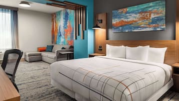 A modern hotel room with a large bed, a desk, a chair, and a colorful abstract painting on the wall.