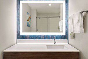 Une salle de bain avec un lavabo blanc, un grand miroir et une douche visible dans le reflet.