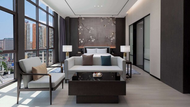 Une chambre d’hôtel moderne, dotée d’un grand lit, d’un canapé et d’une table basse. La chambre dispose de grandes fenêtres offrant une vue sur la ville.