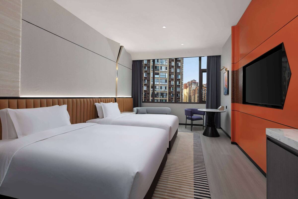 Ein Hotelzimmer mit zwei Betten, einem Fernseher, einem kleinen Tisch und einem Fenster mit Blick auf die Stadt.