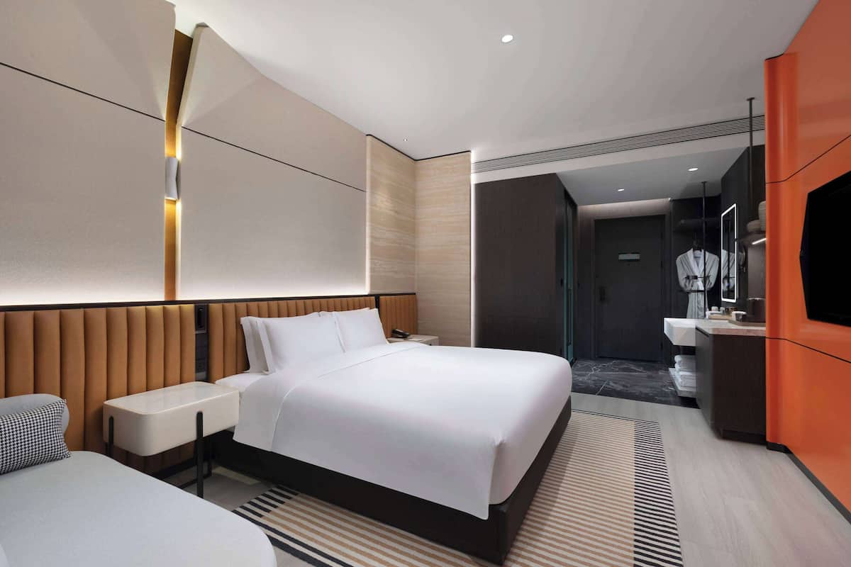 Ein modernes Hotelzimmer mit einem großen Bett, Nachttischen, einem Fernseher und einem durchgegangene Tür erkennbaren Badezimmer.