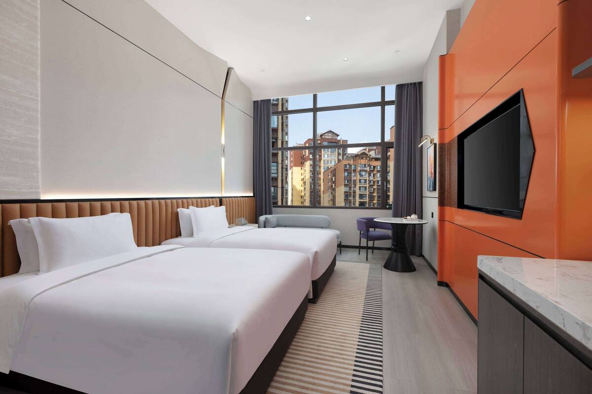 Ein Hotelzimmer mit zwei Betten, einem Fernseher, einem kleinen Tisch mit Stühlen und einem großen Fenster mit Blick auf die Stadt.