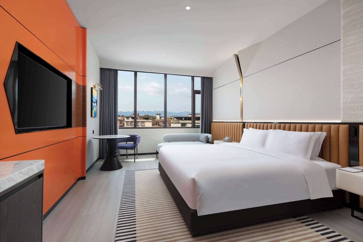 Ein modernes Hotelzimmer mit einem großen Bett, einem Flachbildfernseher, einem Esstisch und Blick auf die Stadtlandschaft.