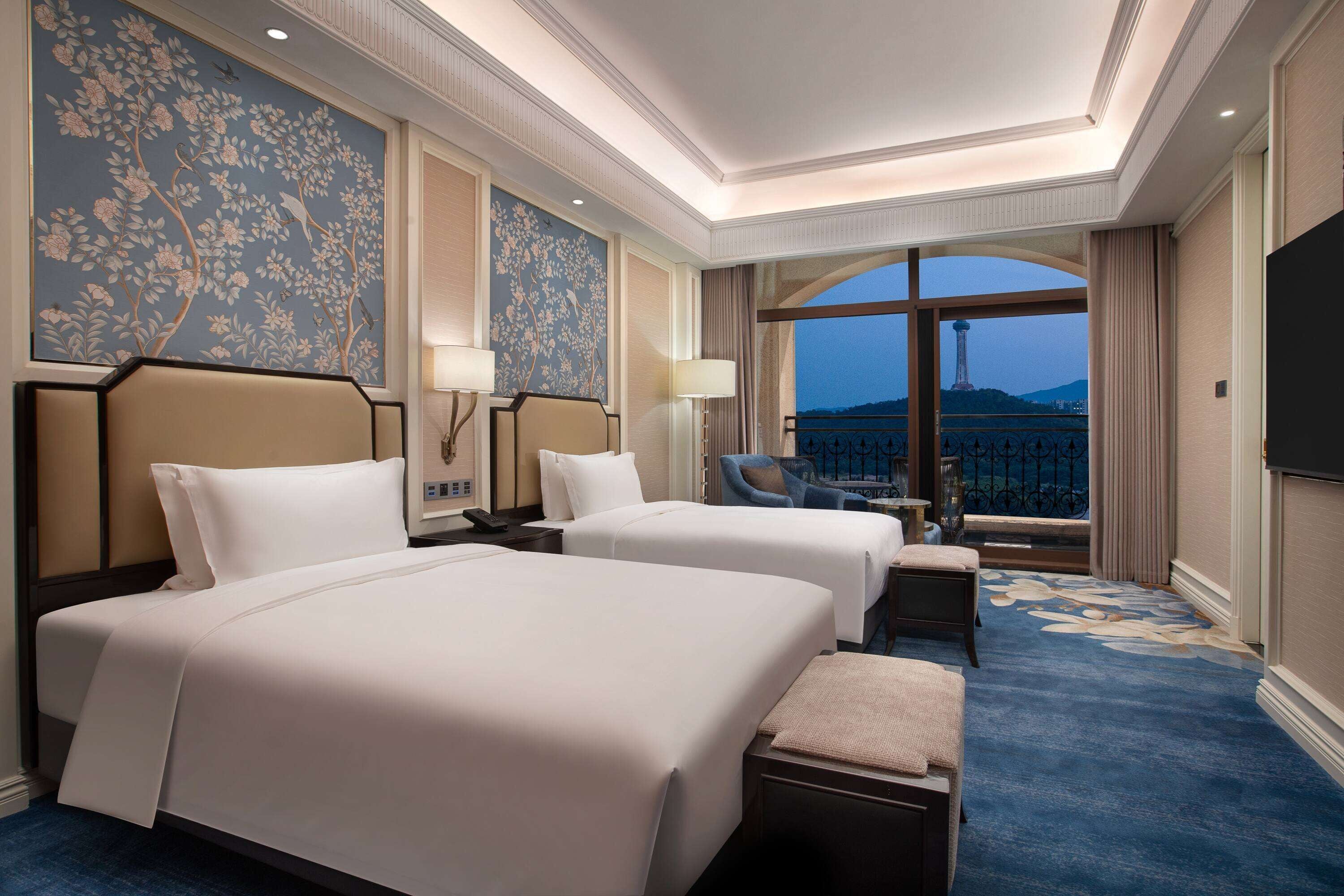 A hotel room with two beds, a desk, a chair, and a large window with a view of a tower.