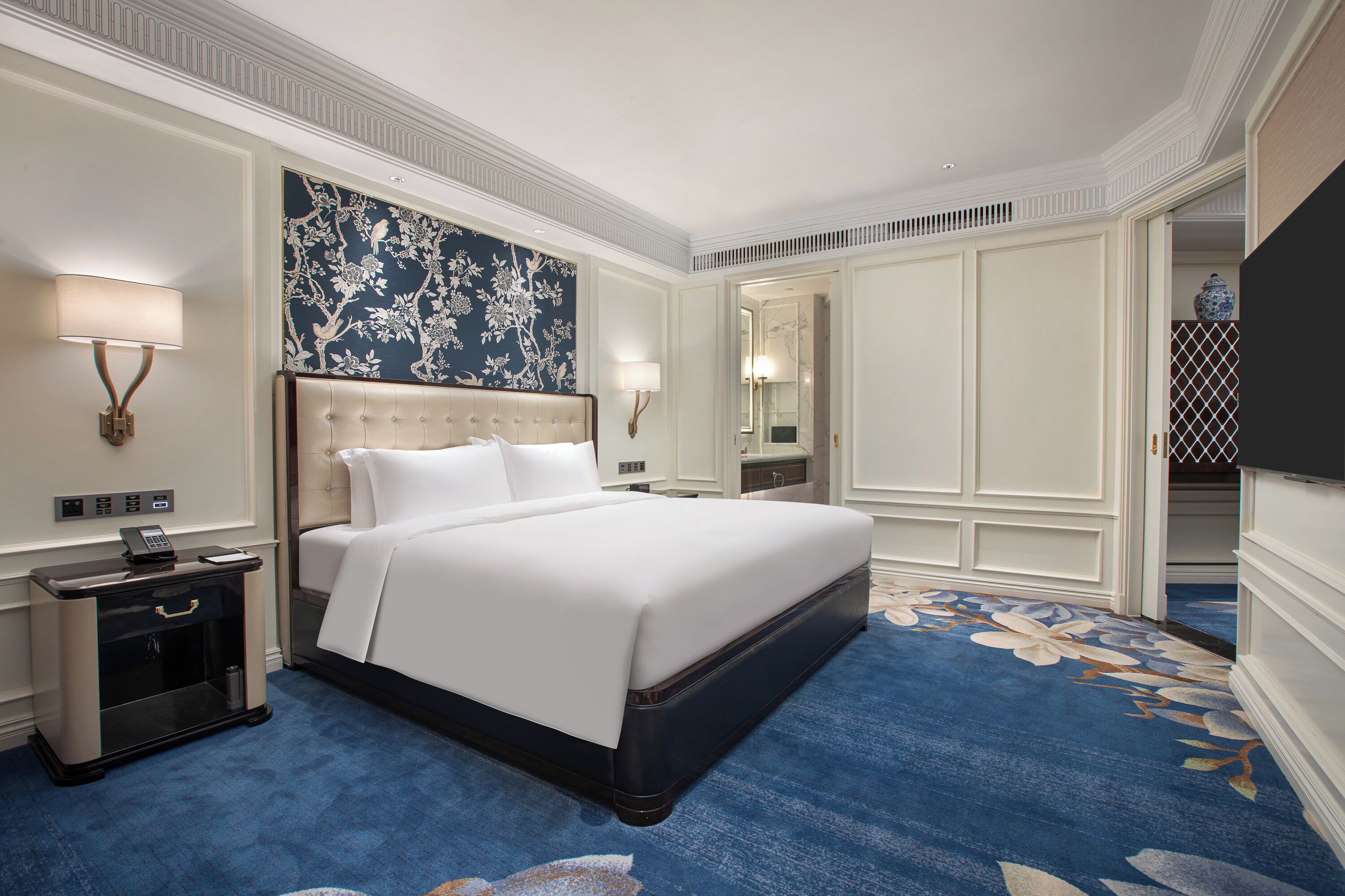 Habitación de hotel con una cama grande, una mesita de noche con lámpara, un espejo en la pared y una alfombra estampada con motivos florales.