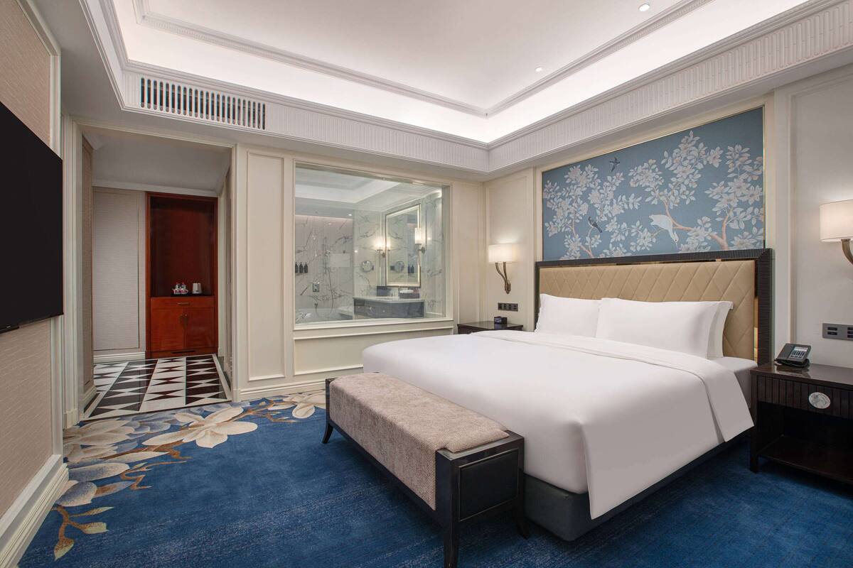 Une chambre d’hôtel avec un grand lit, une banquette, une table de chevet avec une lampe, un miroir et un mur orné d’un tableau représentant des motifs floraux.