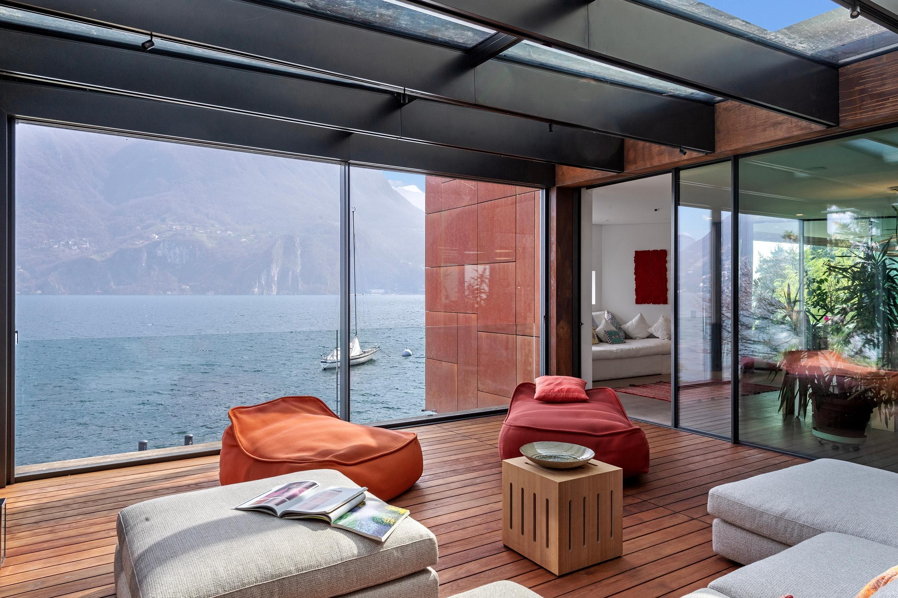 A modern interior with a large glass window overlooking a body of water and mountains. There are wooden floors, a red sofa, and a white couch.