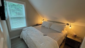 Un dormitorio con techo inclinado, una cama con ropa de cama blanca, dos lámparas de noche y una ventana con persianas.