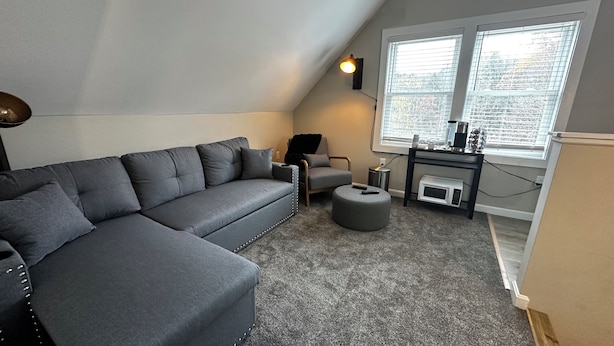 Un dormitorio tipo loft acogedor con un sofá seccional gris, un otoman pequeño y redondo, una silla, una mesita auxiliar, una lámpara y una ventana con persianas.