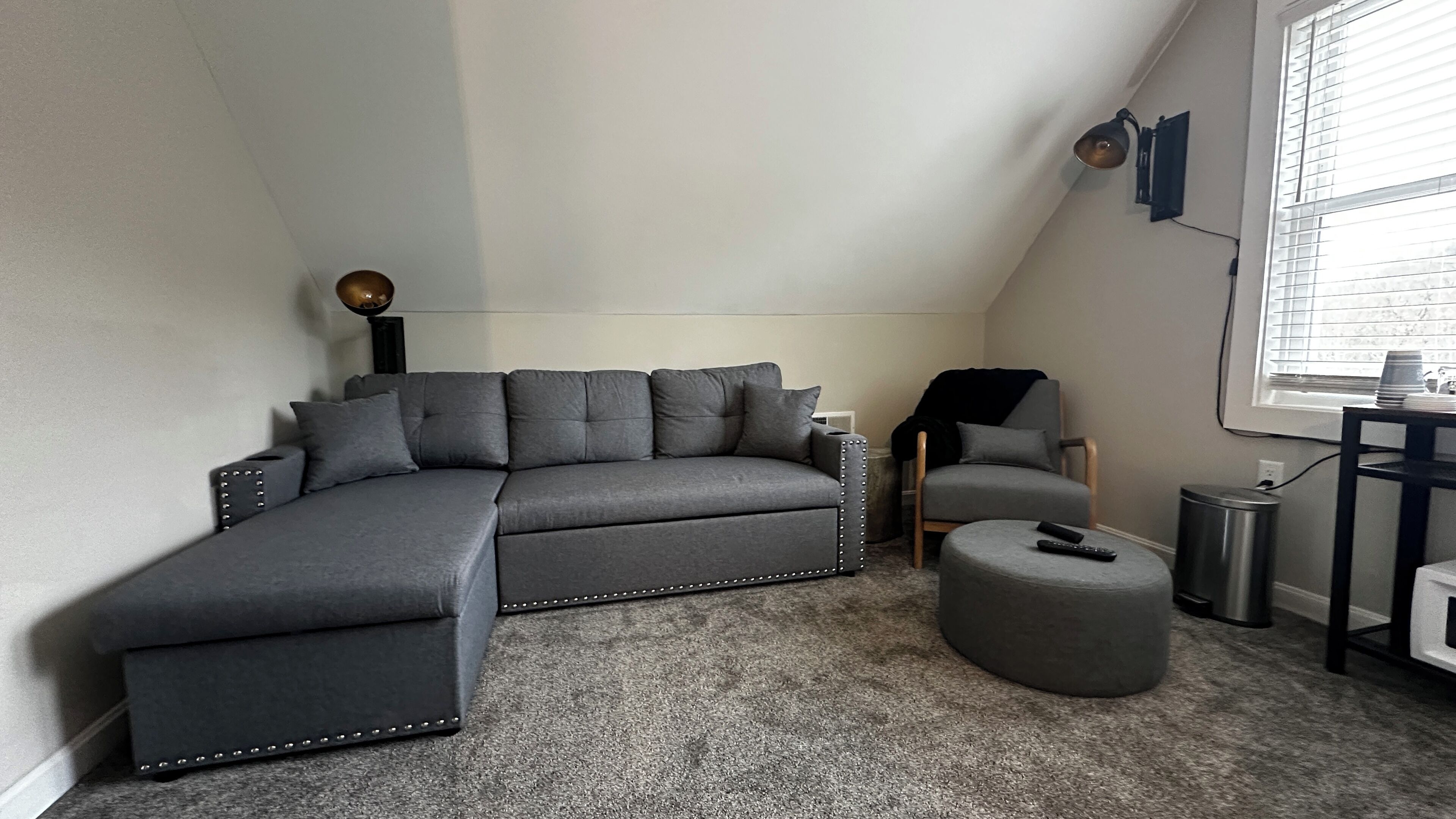 A modern loft bedroom with a grey sectional sofa, a small round ottoman, a grey armchair, and a floor lamp.