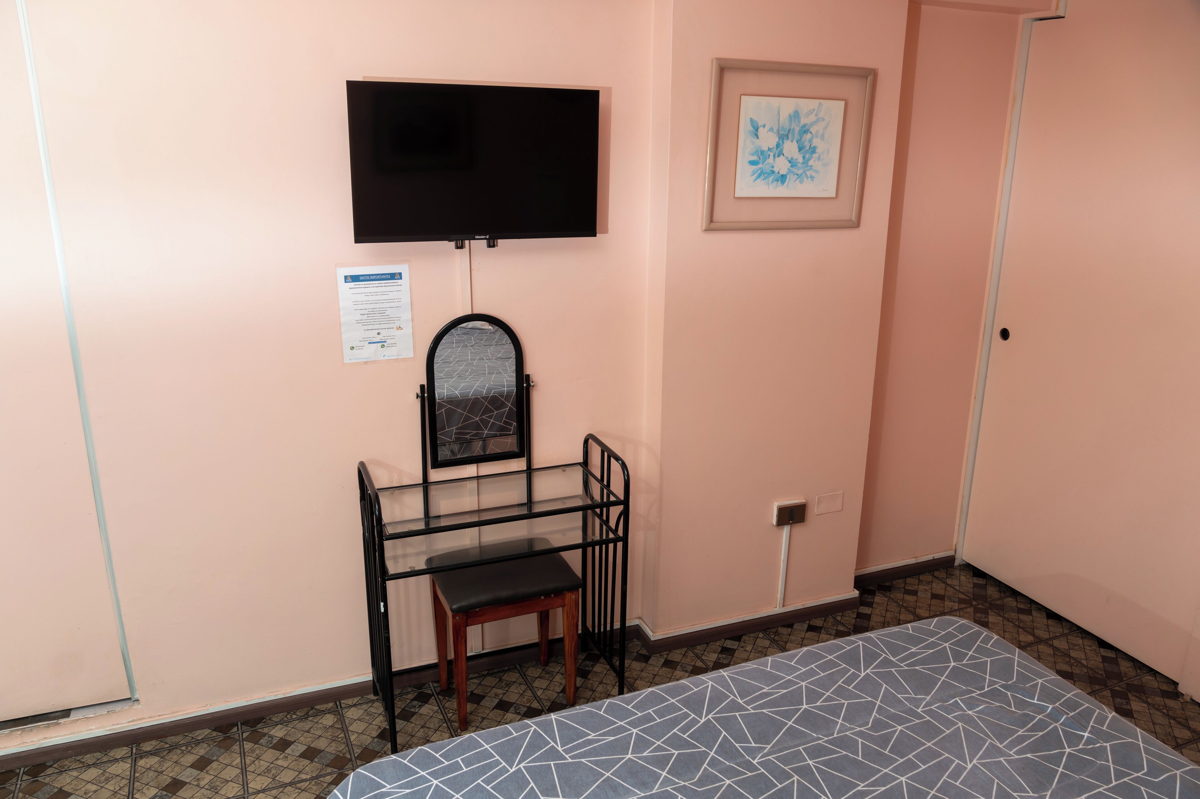 Ein Zimmer mit einem Bett, einem Schreibtisch mit Stuhl, einem an der Wand befestigten Fernseher und einem gerahmten Bild.