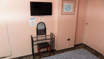 Ein Zimmer mit einem Bett, einem Schreibtisch mit Stuhl, einem an der Wand befestigten Fernseher und einem gerahmten Bild.
