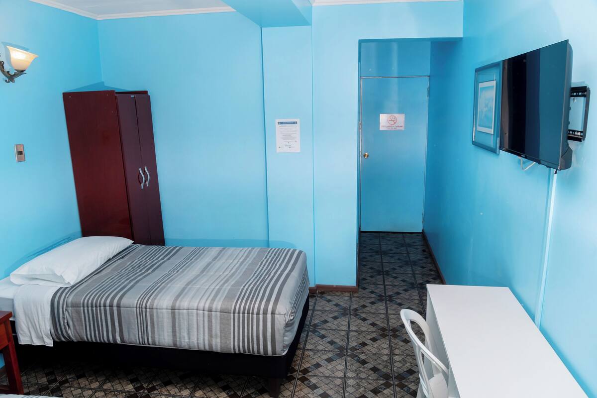 Une petite pièce aux couleurs vives, équipée d’un lit, d’un bureau, d’une armoire et d’un téléviseur fixé au mur.