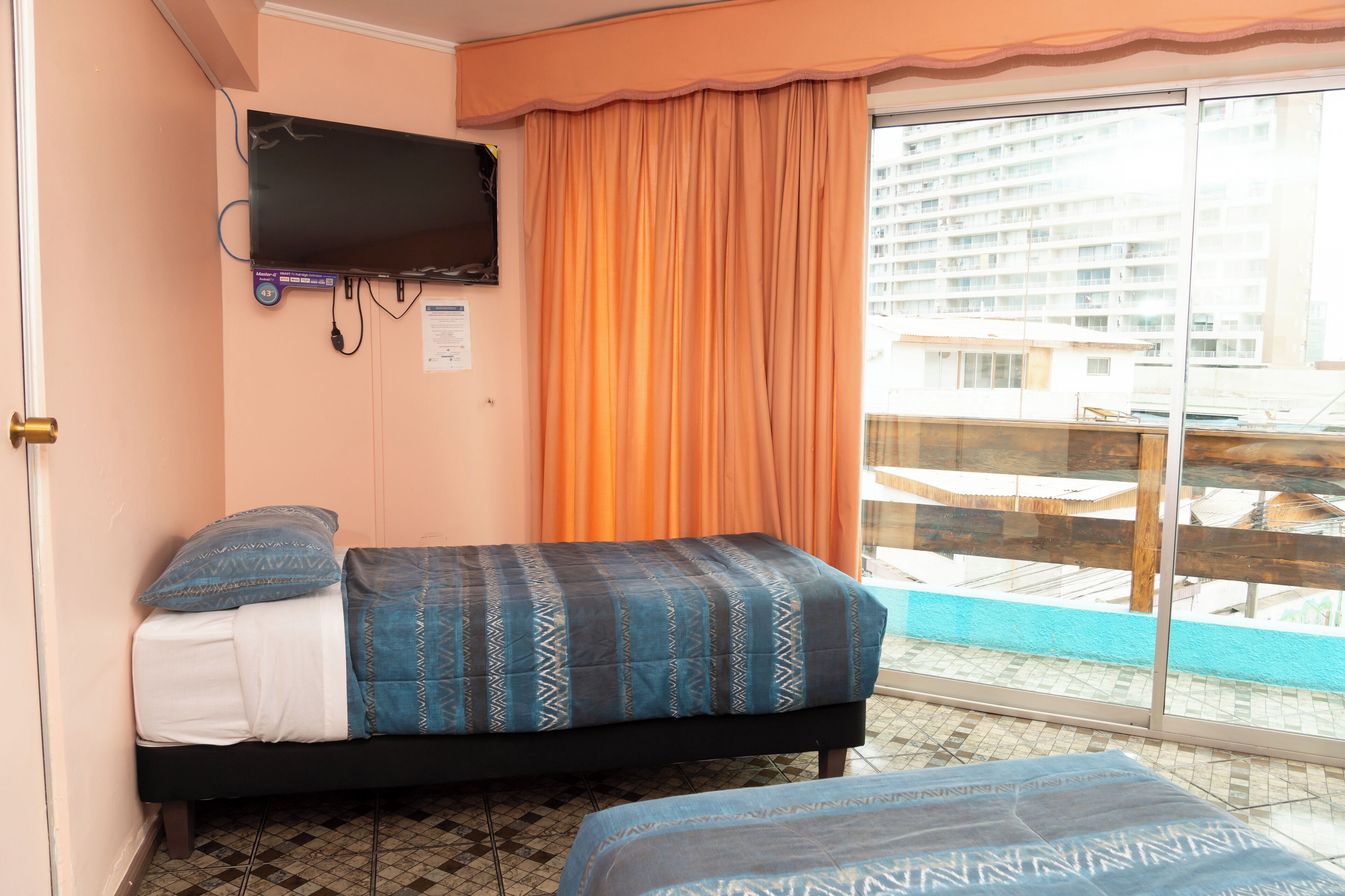 Une petite pièce avec un lit, un téléviseur fixé au mur et une vue sur un immeuble par une fenêtre ornée de rideaux orange.