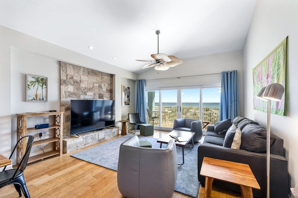 A modern living room with a flat-screen TV, comfortable seating, a ceiling fan, and a large window offering a view of the outdoors. - Beautiful, 2 Bed/2 Bath Pet Friendly Gulf Front Townhome at Portside Villas! (Pensacola Beach)