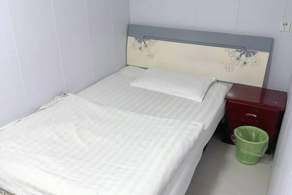 Ein Einzelbett mit schlichtem weißen Bettbezug und Kissen, ein Nachttisch aus Holz mit roter Schublade sowie ein grüner Mülleimer.