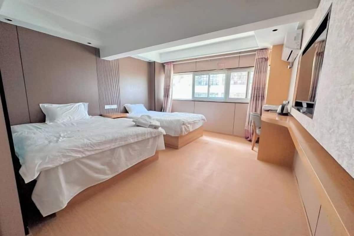 Ein Hotelzimmer mit zwei Betten, einem kleinen Schreibtisch, einem Fenster mit Vorhängen und einem Flachbildfernseher.