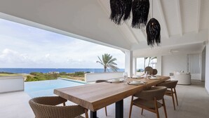 Uma sala de jantar moderna com mesa de madeira e cadeiras de vime, com vista para uma piscina e o oceano.