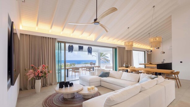 Uma sala de estar moderna com um sofá seccional grande, uma mesa de centro redonda e um ventilador de teto. Há uma área de jantar com uma mesa e cadeiras de madeira, e uma grande janela com vista para o mar.